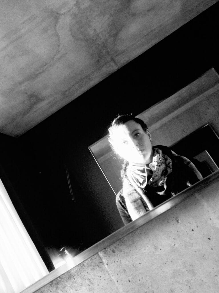Black-and-white portrait of the photographer in a mirror. The high-contrast light cuts er face in two, with an overexposed side on the left and a shadowed side on the right. The composition is very graphic, with parallels tilted to the right. The ceiling is made of concrete, and the atmosphere is sombre, with the room plunged into half-light, the harsh light cast by the window on the right and the inexpressiveness of the face.