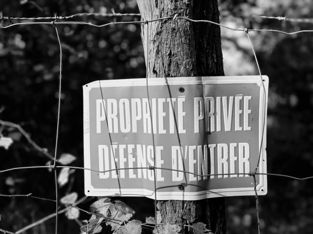 Close-up of a ‘Private property. No trespassing’ sign nailed to a fence post, behind a fence with barbed wire added, welcome...
