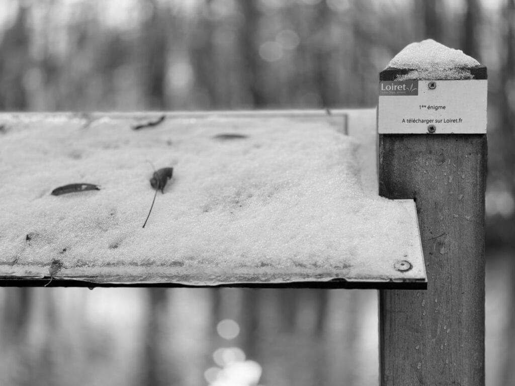 View of an educational panel along a hiking trail. The sloping main panel is covered in snow. The situation sticker on the post reads 'First puzzle'. Indeed.
