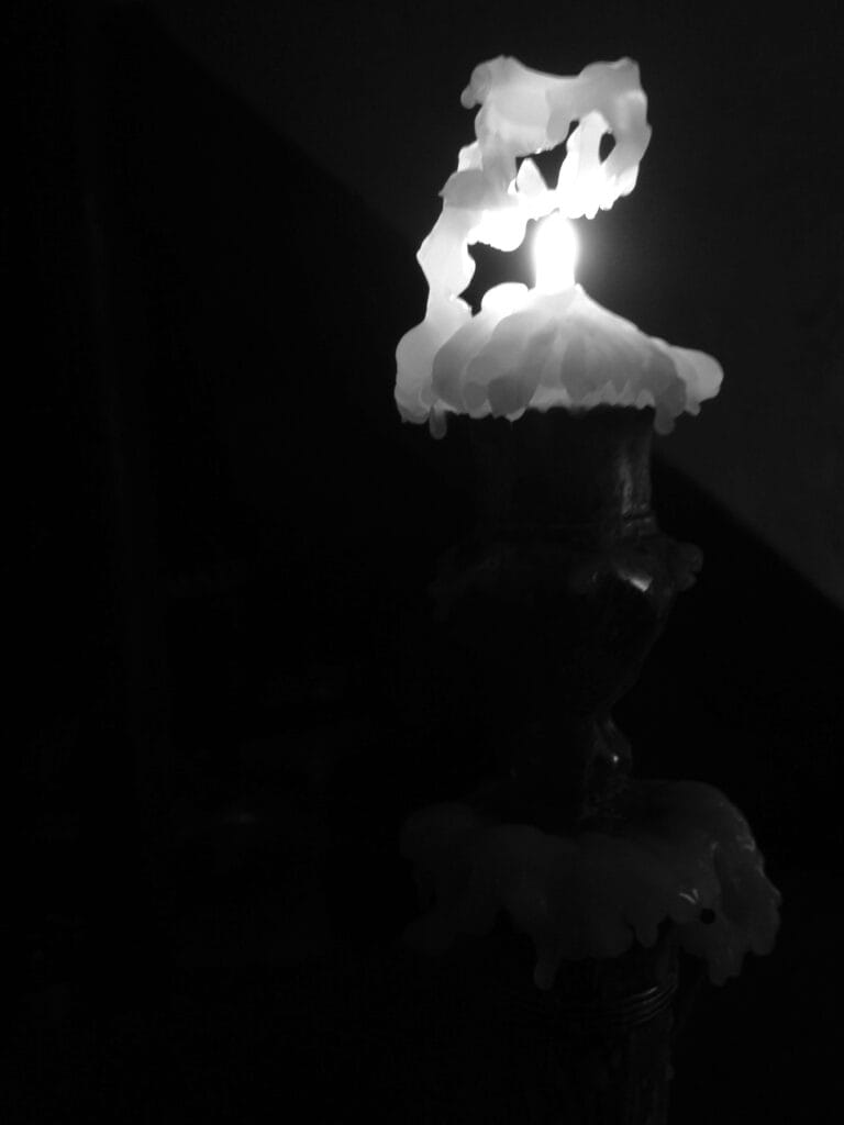 A view of a candle in a metal candlestick, lit only by the flame of the candle itself. The candle has burned down considerably, and the melted wax has formed a kind of gantry above the flame.