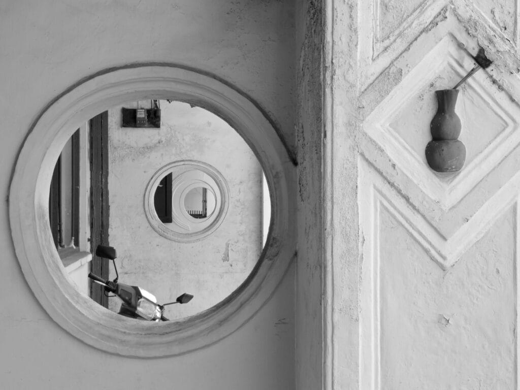A detail of the party wall of a porch, with a round dormer window through which the porch wall of the next-door house can be seen. That house has a similar dormer window through which the porch wall of the next-door house can be seen. That house has a similar dormer window through which the porch wall of the next-door house can be seen. …
