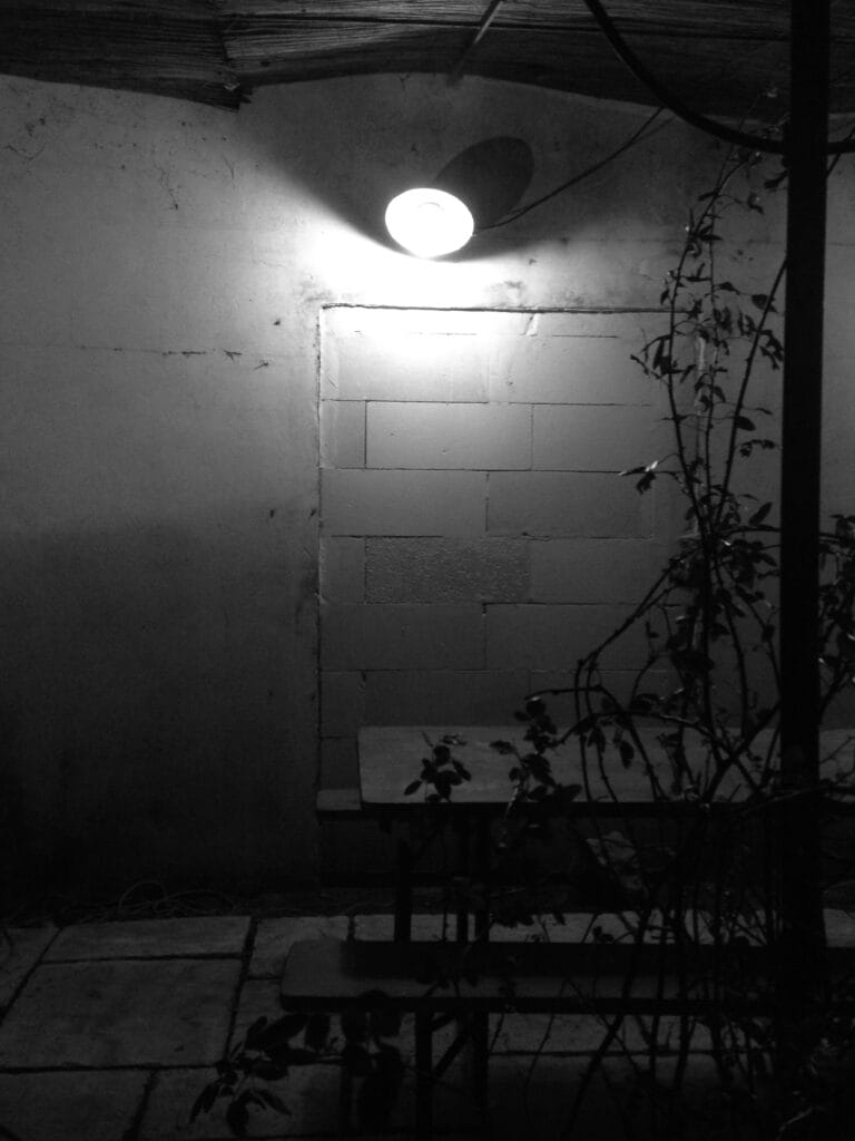 A courtyard with a picnic table, lit by a lamp fixed to a wall where an old opening has been filled in with breeze blocks.