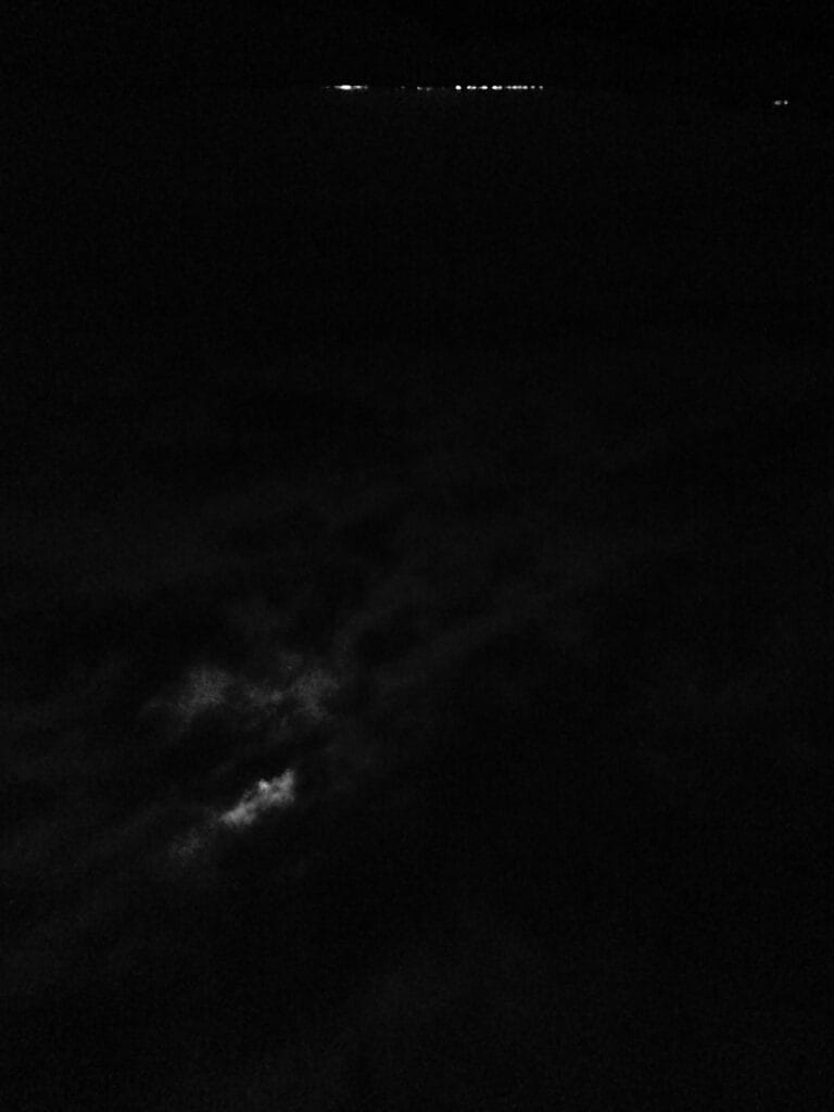 Landscape of a night sky with the moon peeking through the clouds, and the lights of a village twinkling on the horizon.