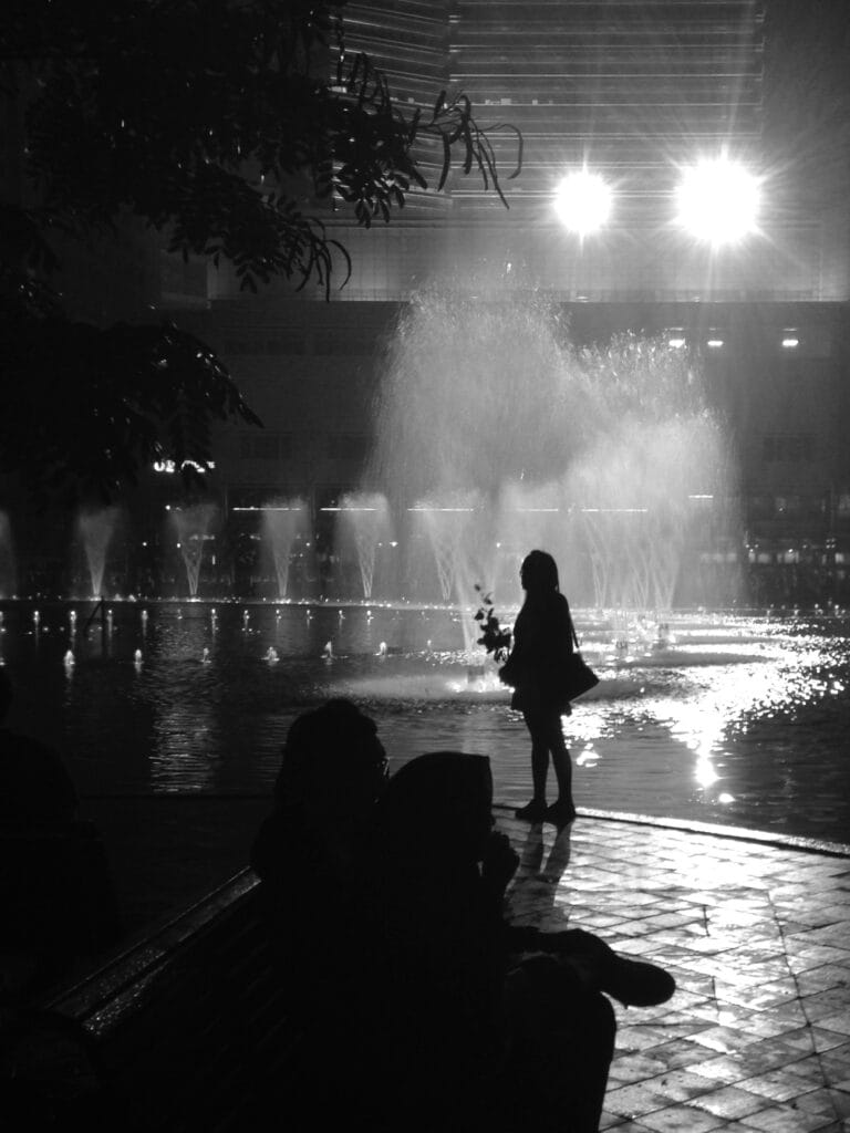 A person stands alone, holding a rose, in front of a pool of water jets in an urban square. The image is taken against the light in front of large spotlights, so we can only see er silhouette.