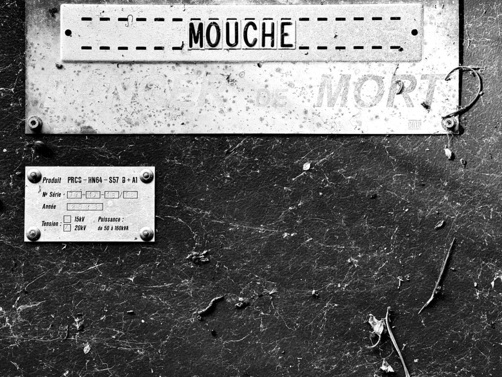 French energy supplier sign on a transformer with the location name 'Mouche,' which means 'A fly', and slightly faded underneath 'Danger de mort', which means 'Danger of death'.