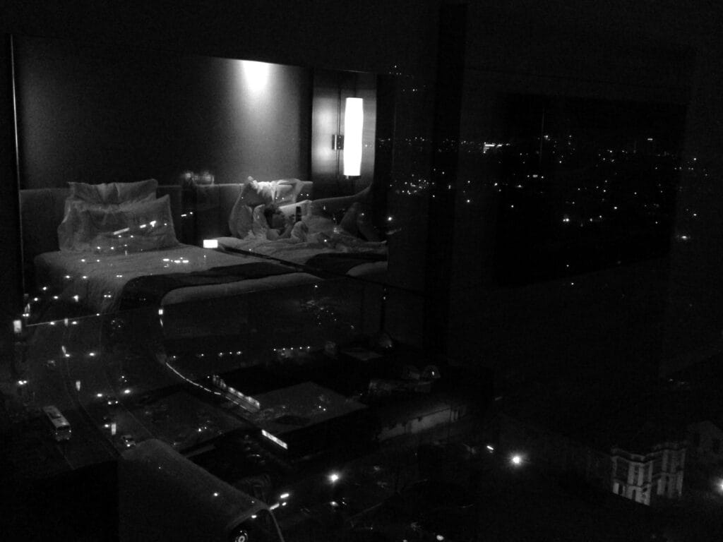 A hotel room's bay window reflects a person reading in bed against a backdrop of the city by night.