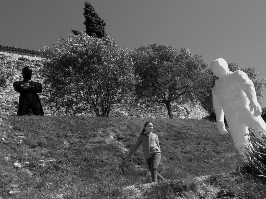 This is a view of a terraced Mediterranean garden with olive and cypress trees, as well as an art installation. Two statues of men in threatening poses are visible in the image: one black on the upper terrace and one white on the lower terrace. A nonchalant little girl walks by on the path.
