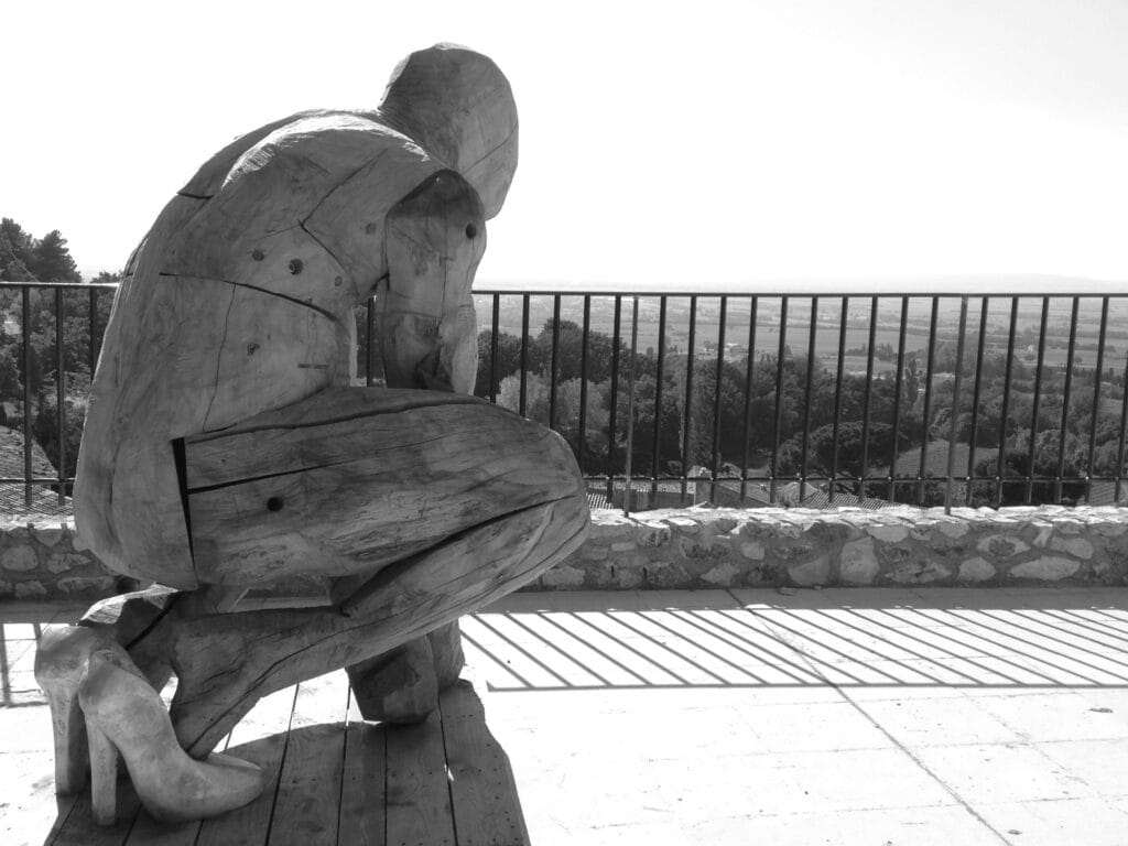 A wooden statue in high heels is seen from behind as e looks down at the plain below. Squatting on the high terrace, e appears ready to bounce.