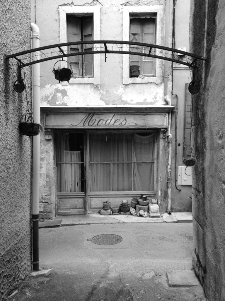 This is a view of the front of an old store with a painted wooden sign that reads 'Modes.' The facade is dilapidated, and faded, old curtains hang behind the checkered windows.