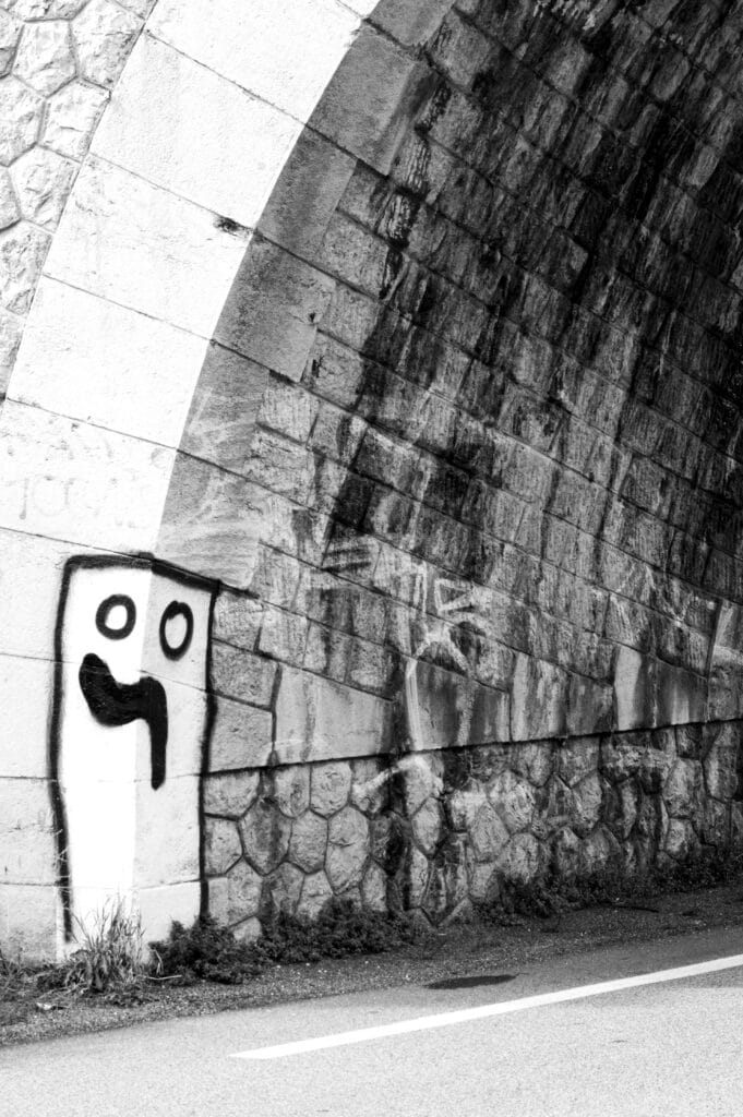 View of a graffiti depicting a bollard-shaped figure with two pupil-less eyes and a sagging mouth, painted on the cornerstones of a railway bridge over a road.