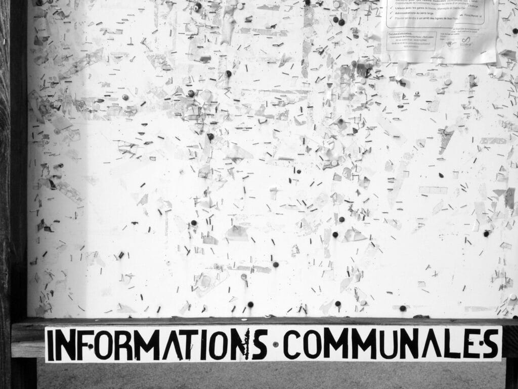 The municipal notice board is almost empty, with just the tip of a poster visible, but it is massively overgrown with staples from previous postings. The title 'Informations communales' (municipal informations) can also be seen.