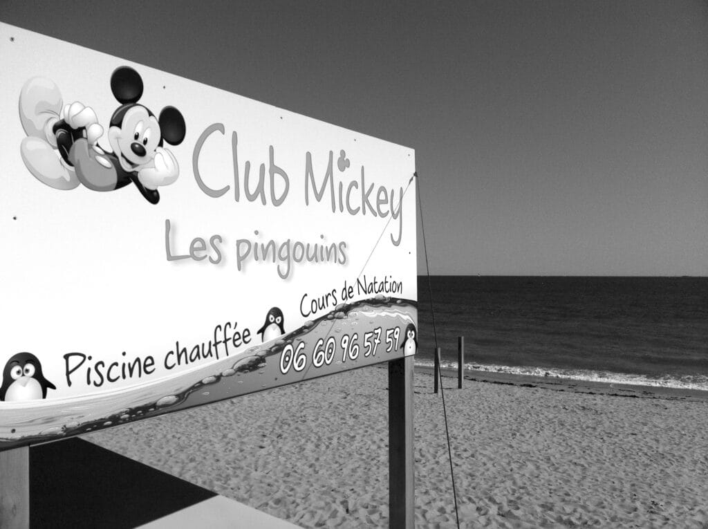 View of the sea with a large Mickey Club 'The Penguins' billboard in the foreground. 'Clubs Mickey' are the larger franchise of 'clubs de plage' in France, which are not 'beach clubs' but kind of mini-camps on the beach where you can leave your kid to play for a couple of hours. (I couldn’t find how it is called elsewhere…??)