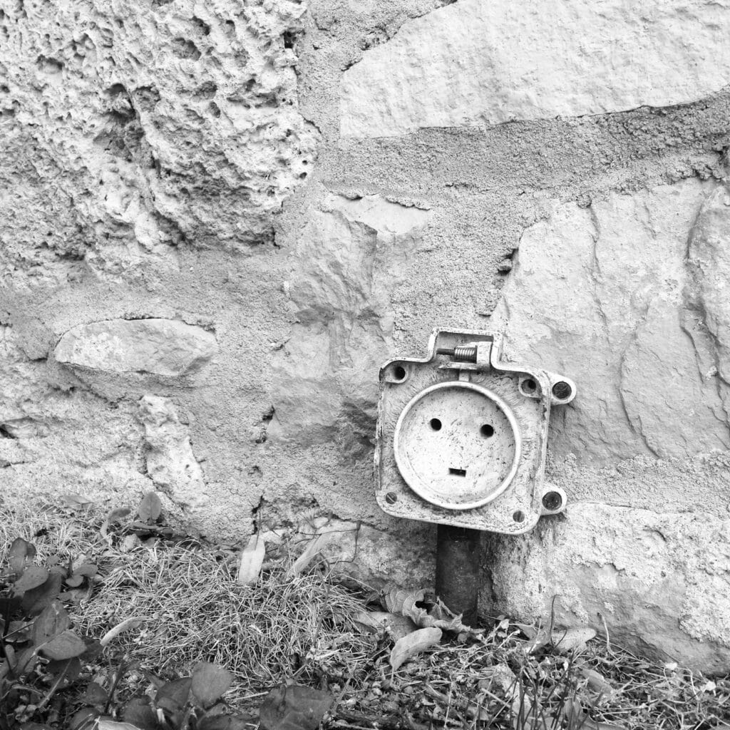 An outdoor socket in the shape of a square and a piece of cable emerge from the ground between the lawn and a wall. Without its protective cover, the plugs inscribed in a circle resemble a face, making the whole thing look like a small robot.
