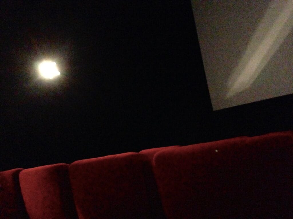 In a movie theater, rows of seats shot from behind, with the screen in the background.