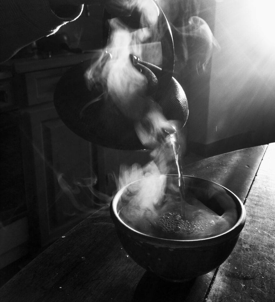A hand pours hot tea from a cast-iron teapot into a stoneware bowl. The rising sun's low-angled rays enhance the curls of steam.