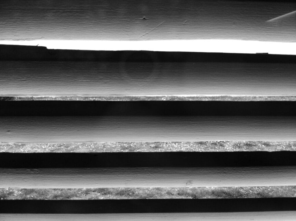 Close-up on the slats of a shutter. Outside the white light of a hot day.