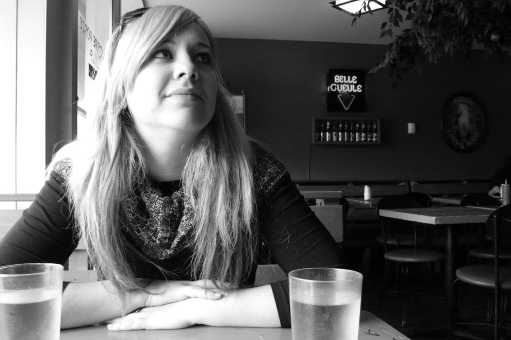Portrait of a person in a bar, seated in front of drinks, er face highlighted by the beautiful light coming from the bay window on the left of the picture. The wall in the background features a row of beer bottles and a neon sign for the 'Belle Gueule' ('beautiful face') brewery.