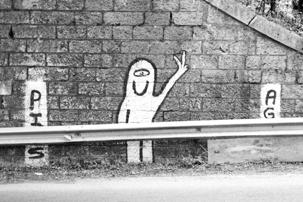 Graffiti along the road, on the wall at the entrance to a tunnel and behind the guardrail. The graffiti depicts a ghost-like little person with one eye, a big smile, and making a victory sign.