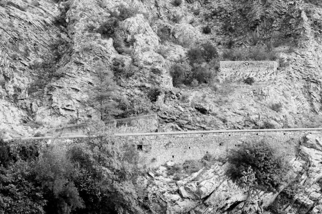 An eye-shaped graffiti appears on a retaining wall above a road on a mountainside.