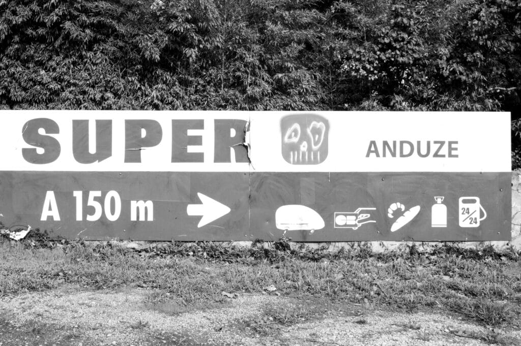Sign indicating a ‘Super U’ chain supermarket 150 metres away. An inspired artist has transformed the ‘U’ into a skull graffiti. The sign features pictograms for the different services: a car that looks like a rock on wheels to symbolise the click & collect, a card which reads 'C.B.' (for 'carte bancaire' in French, i.e. bank card), bread and croissants that look like small bones for the bakery, a gas bottle and a 24/7 petrol pump.