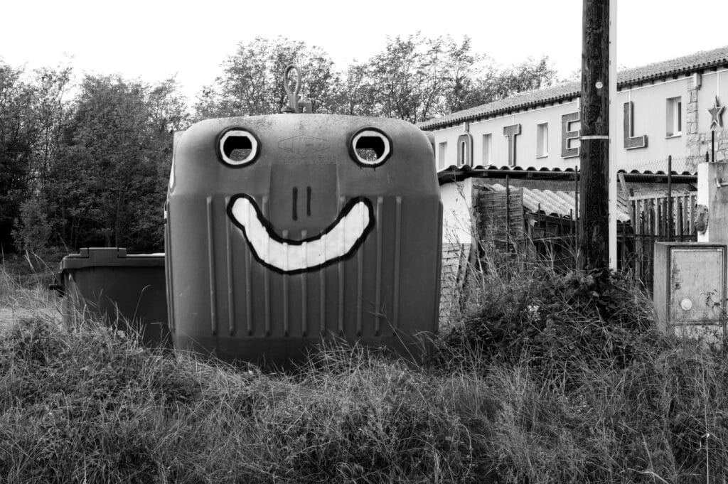 A street artist transformed a glass recycling bin into a face by painting white circles around the holes to create eyes and drawing a big smile underneath.