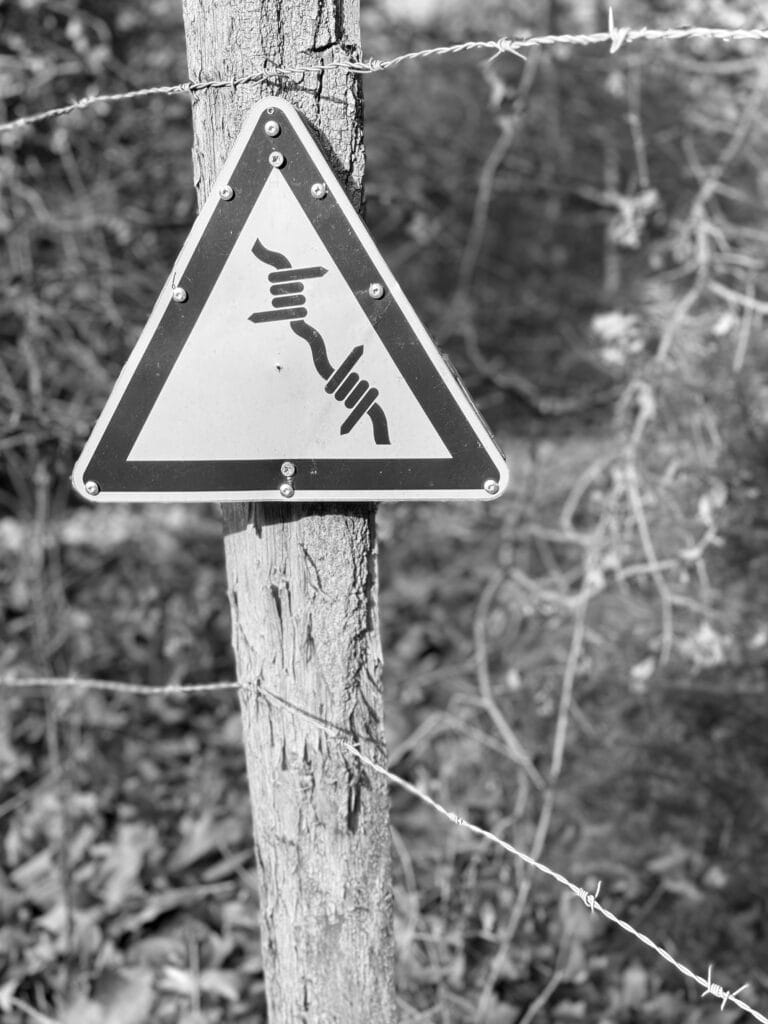 Sign reading ‘Beware of barbed wire’ fixed to a fence post with no fewer than ten nails.