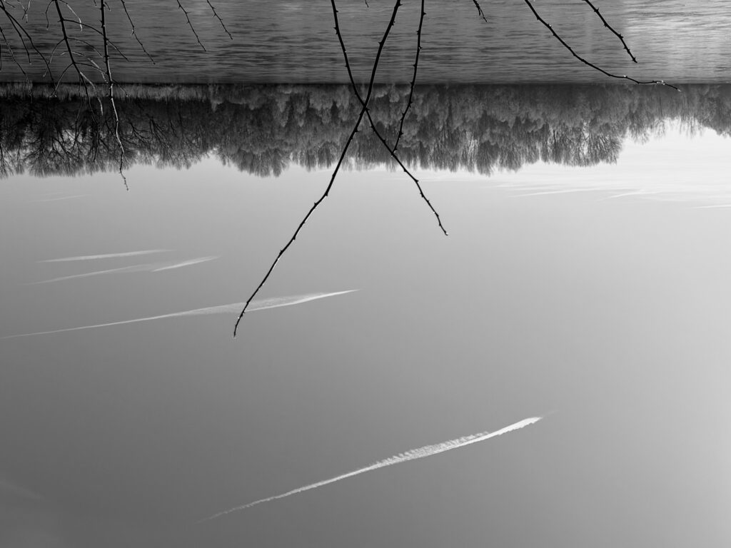 A view of a river in winter under a vast blue sky. Contrails in the sky. The opposite bank is wooded. The image is upside down.