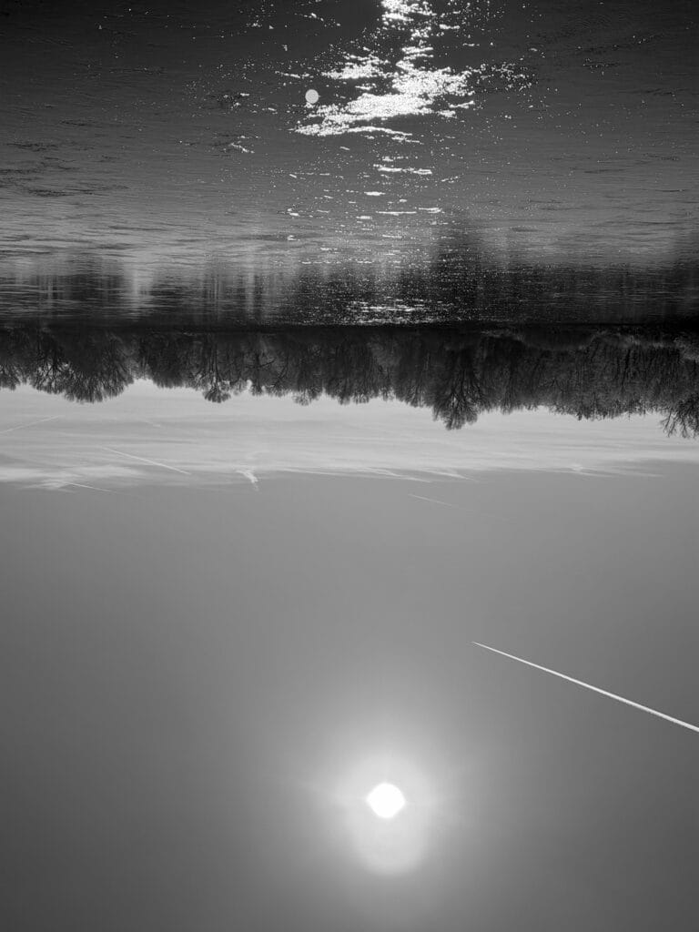 A view of a river in winter, with the sun’s rays reflected on the water. An aeroplane is flying across the sky. The opposite bank is wooded. The image is upside down.