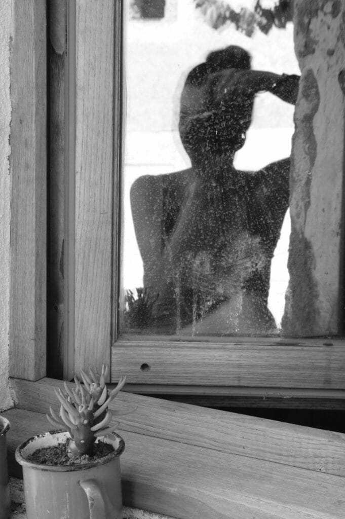 Self-portrait of the photographer in a window pane, with a small cactus placed on the sill.