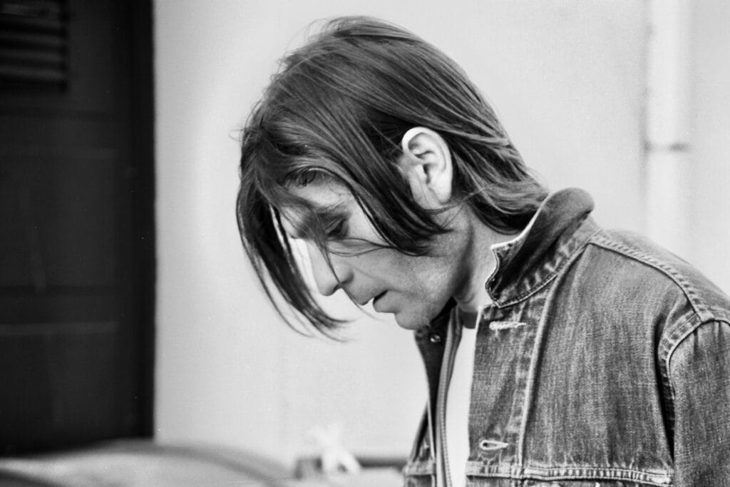 A street portrait of a person wearing a denim jacket, er head tilted forward, framed by soft strands of hair. In the background, the wall of a building and a door.