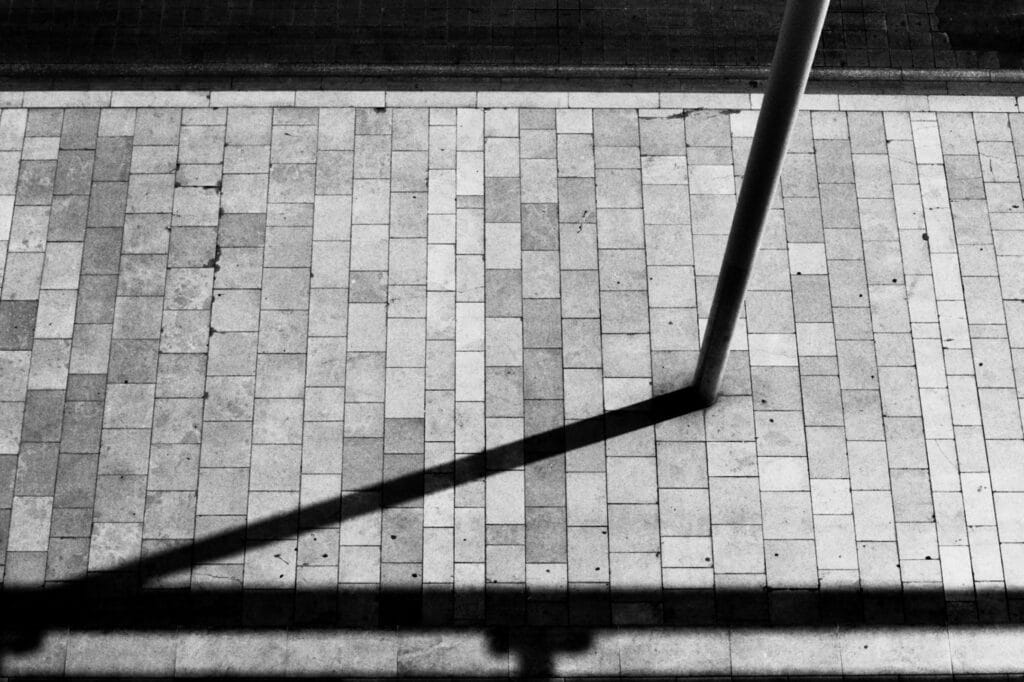 The sharp shadow of a post on the cobbled pavement. Photo taken from above, looking down from the roof terrace of a building.