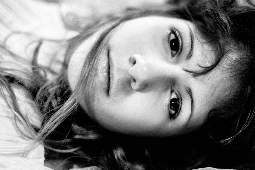 A close-up portrait of a person lying down, with pale skin and tousled strands of hair, staring at the camera with serene, weary eyes.