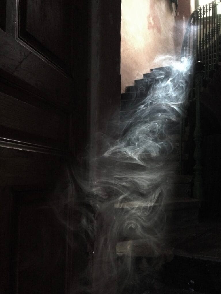 A door opens onto a staircase in the dim light. Smoke appears in the light filtering through the window at the top of the stairs.
