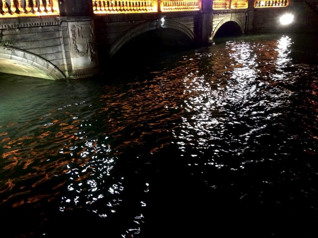 A river beneath a bridge, with nice night-time lighting reflecting on the water.