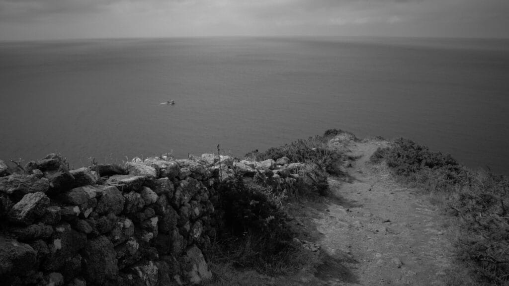 A ridge path overlooking the sea, bordered by a low stone wall, which ends just above the cliff edge. Below and on the horizon lie the sea and a patch of sky.