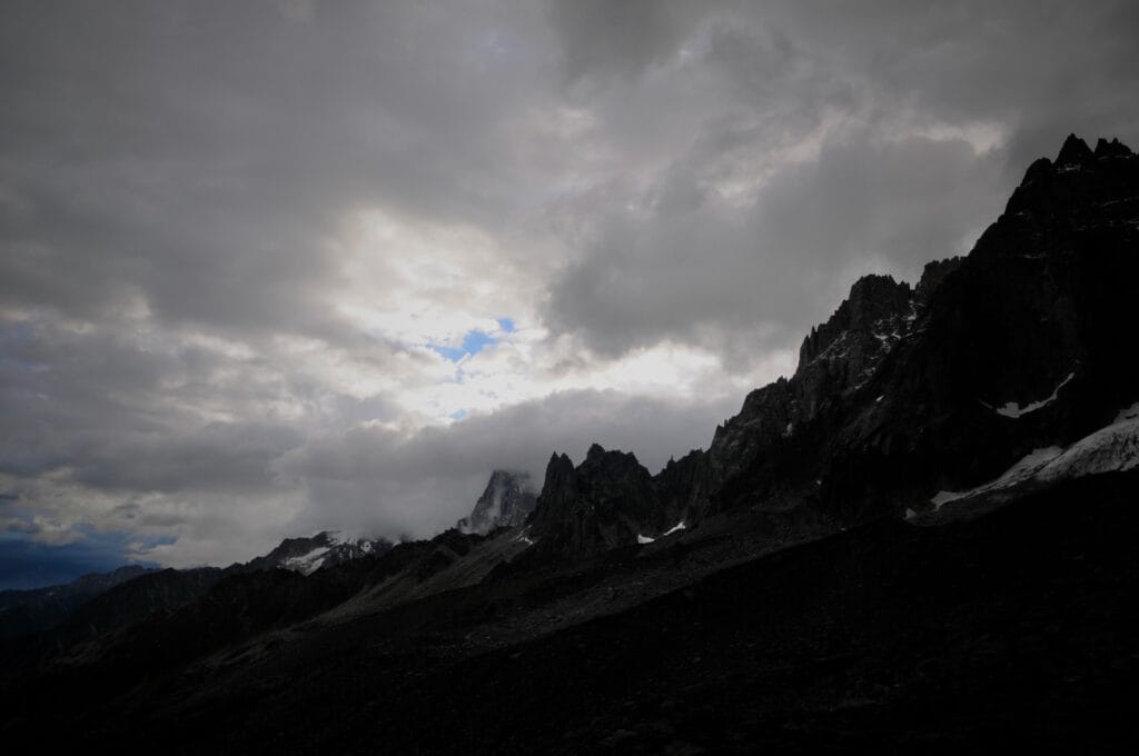 An alpine landscape consisting of a range of very dark rocky peaks beneath a heavy sky, through which two patches of blue sky shine.