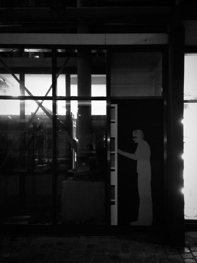 A life-size collage of a masked figure, seen in profile, in white against a black background, on the façade of a glass building, at night.