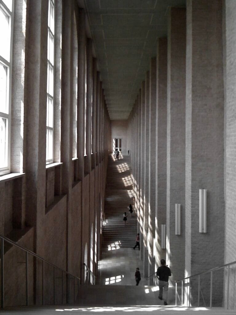 A corridor inside a brick building with two large staircases, lit by tall windows on the left.
