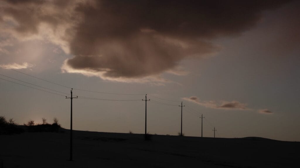 A ridge line with electricity pylons silhouetted against a cloudy sky at sunset.