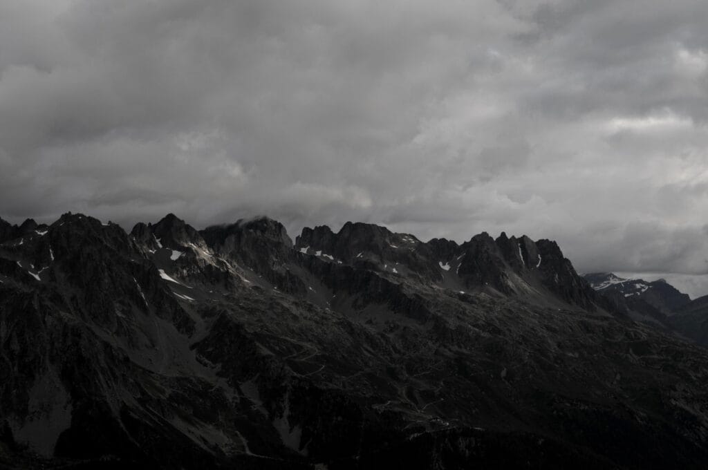 A chain of dark mountain peaks beneath a heavy sky. A single ray of sunlight illuminates a small patch of rock.