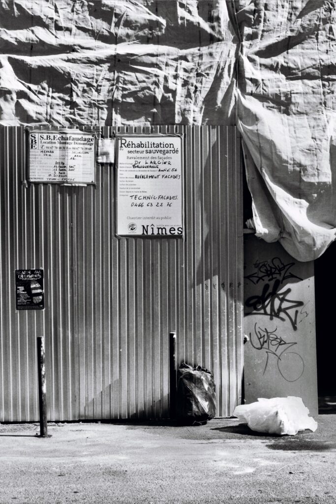 Scaffolding covered with a tarpaulin. A sign reads ‘Renovation of a conservation area’. Rubbish bins have been left on the pavement in front.