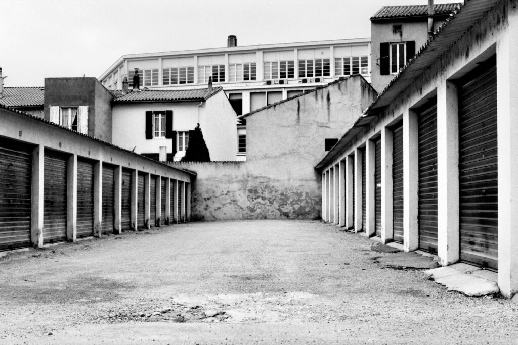 Two rows of garages facing each other, with other buildings in the background. A run-down, 1970s-style atmosphere.