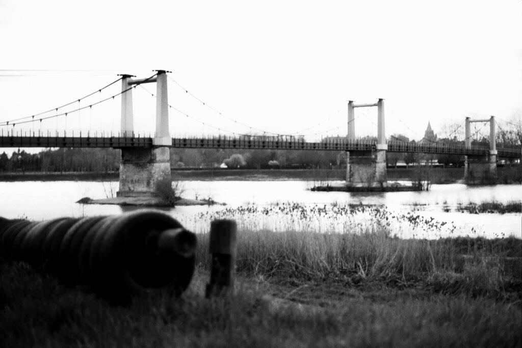 A view of a suspension bridge over a river, taken from the riverbank, with a church spire in the background.