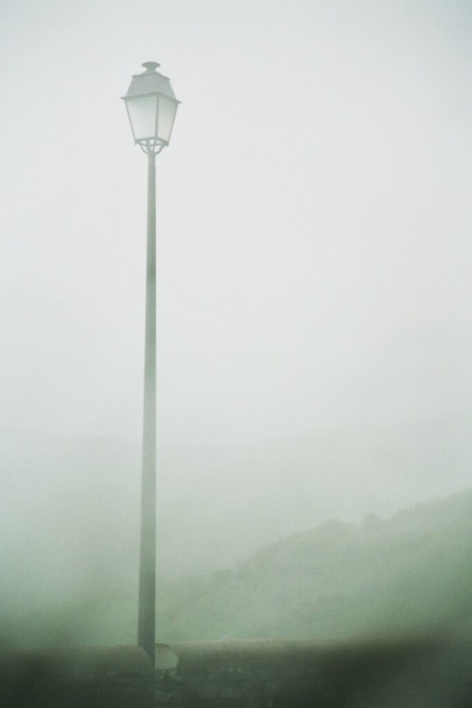 A street lamp and a low wall in the mist. A landscape of wooded hills can be made out in the background.