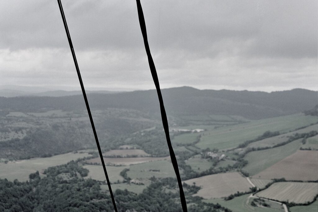 Two vertical power lines cut across a rolling landscape of fields and woods beneath a grey sky.