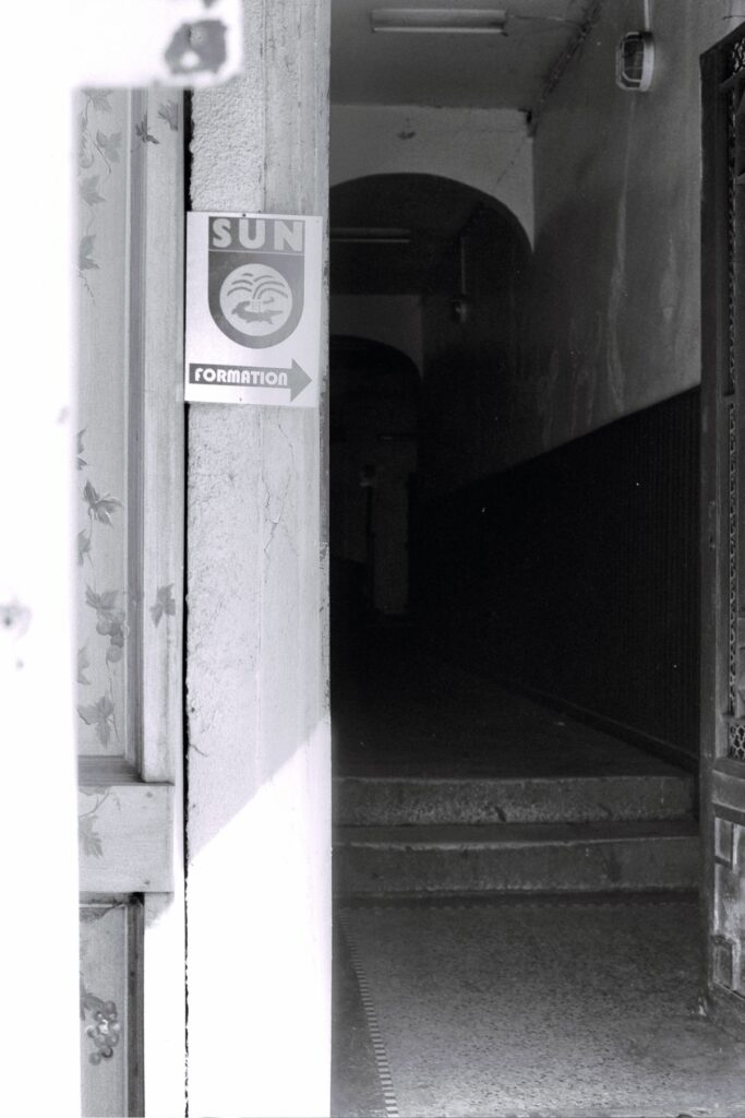 The entrance hall of a block of flats in semi-darkness. A sign with an arrow points to ‘Sun Formation’ (‘Sun School’).