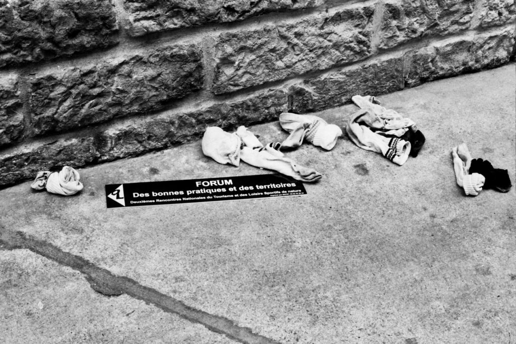 On a pavement alongside a wall, several worn-out socks have been left behind, along with a sign reading in French ‘Forum des bonnes pratiques et des territoires, deuxièmes rencontres nationales du tourisme et des loisirs sportifs de nature’ (‘Forum on Best Practice and Regions: Second National Conference on Tourism and Outdoor Sports’).
