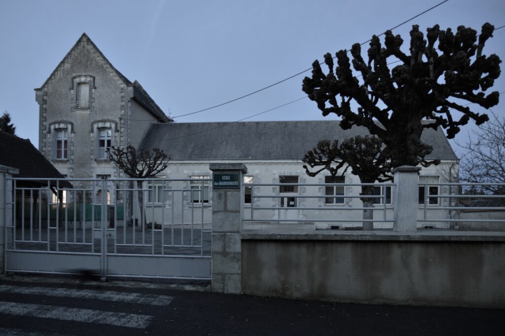 A view of the École des Marronniers (‘Chestnuts school’ in French) at dusk, with the gate in the foreground, followed by the courtyard with its chestnut trees pruned into stumps and the covered playground, and finally the school building.