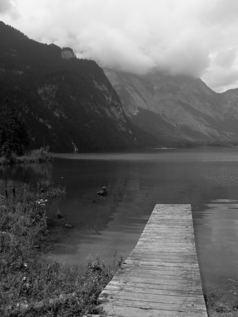 A wooden pontoon jutting out into a lake surrounded by mountains whose peaks are shrouded in clouds.