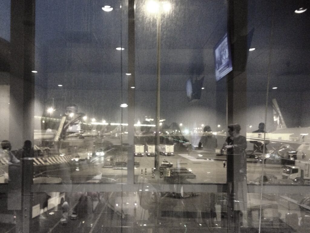 A reflection in the windows of an airport departure lounge at night: reflections of passengers in the glass and the planes parked on the runways.